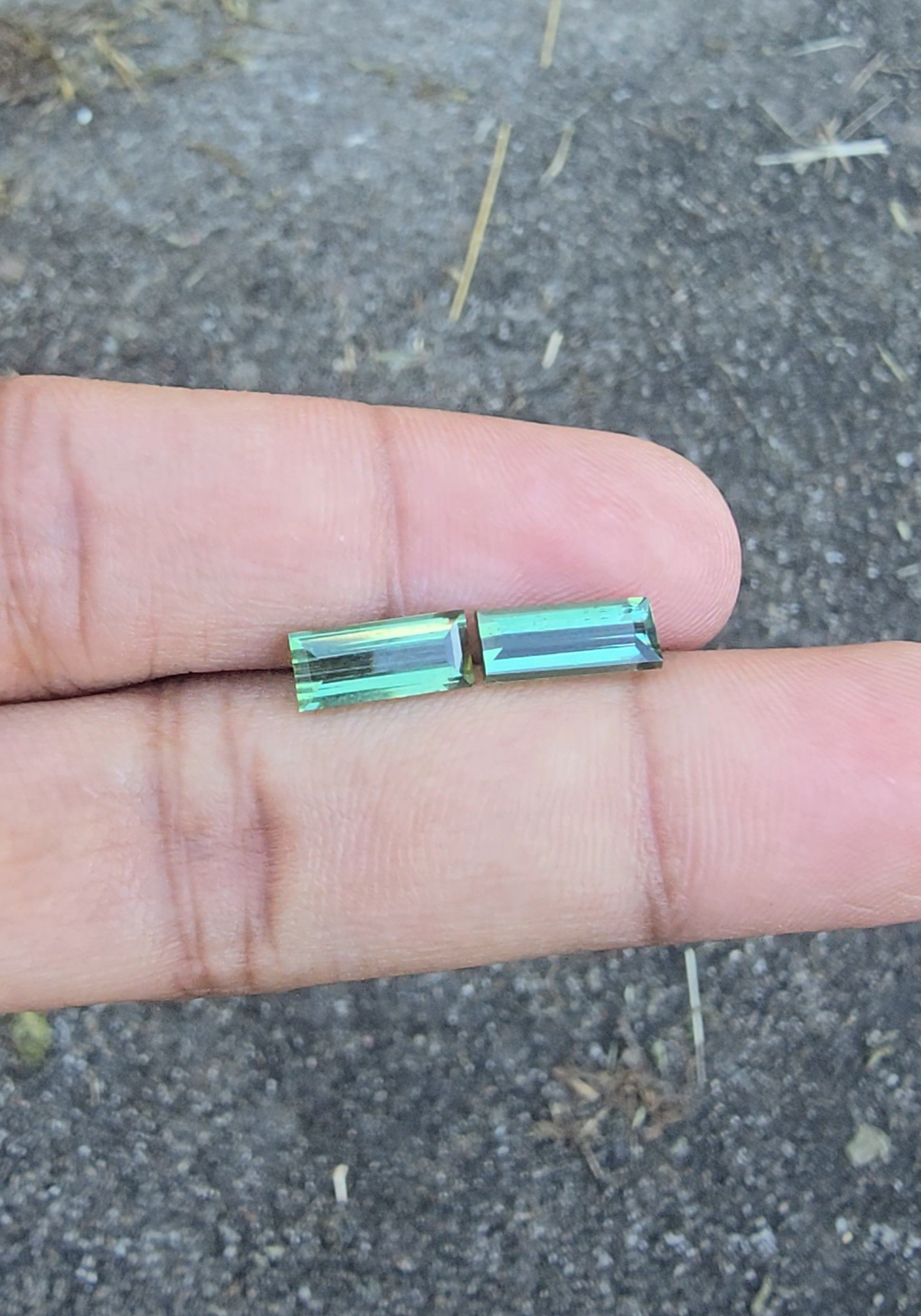 Two small rectangular green gemstone beads held between a person's fingers against an outdoor asphalt backgroun