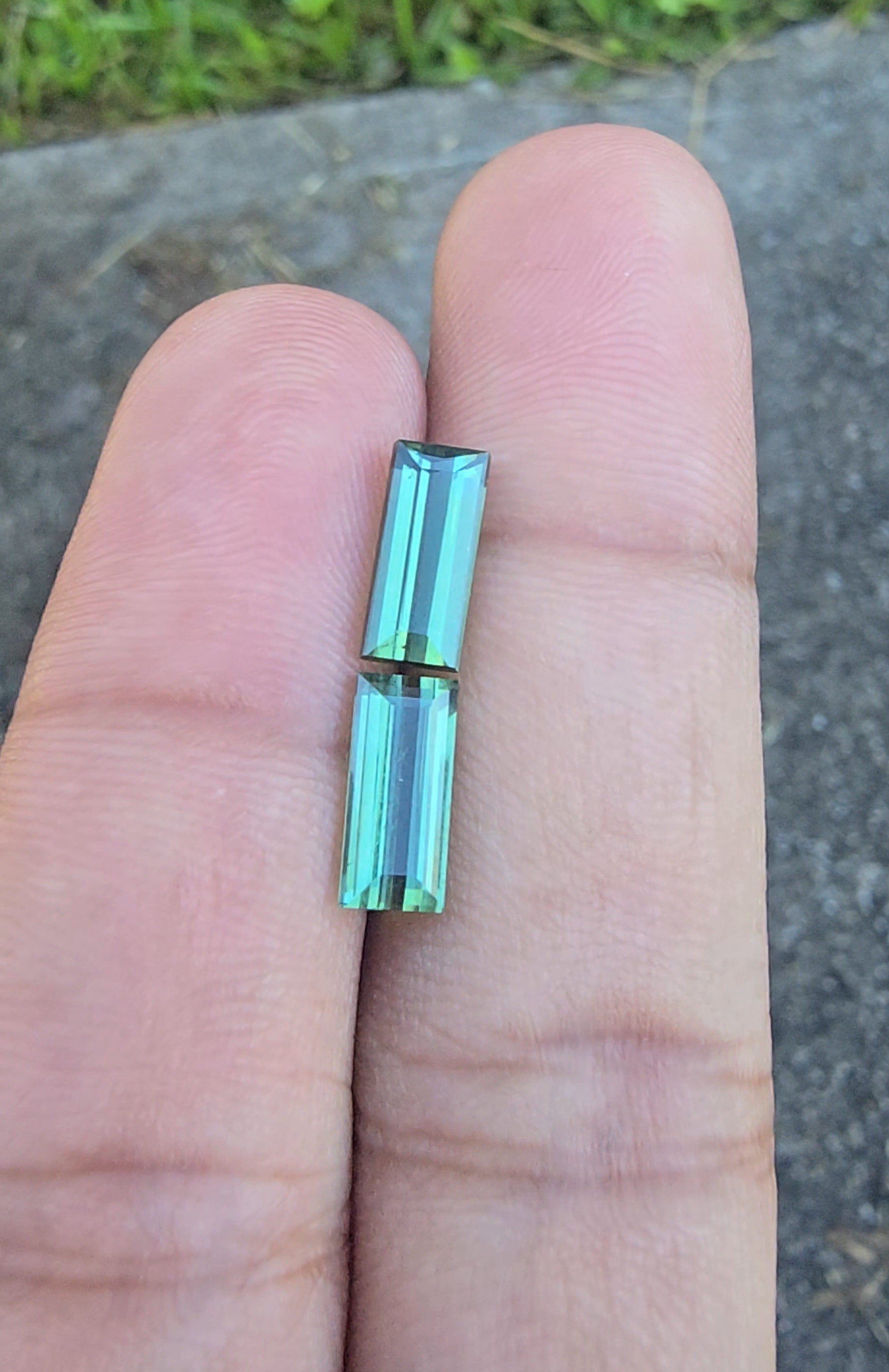 A close-up of a person's fingers holding a small, rectangular, iridescent green gemstone or bead against an outdoor background of pavement and grass.